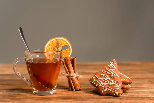 Glass cup of tea with sliced orange near cinnamon sticks and gingerbread cookies isolated on grey — Stock Photo