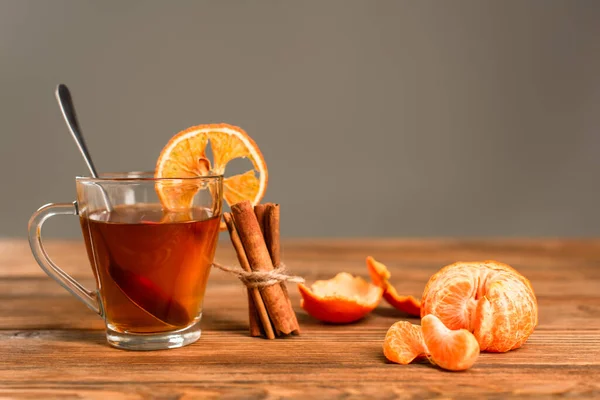 Glass cup of tea with sliced orange near cinnamon sticks and peeled tangerine isolated on grey — Stock Photo