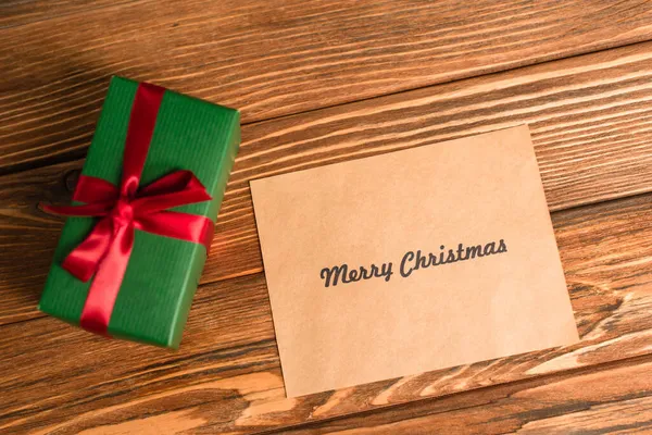 Top view of greeting card with merry christmas lettering near wrapped green present on wooden surface — Stock Photo