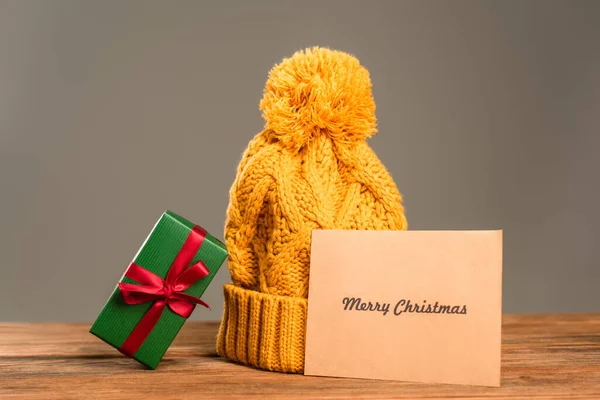 Grußkarte mit frohen Weihnachten Schriftzug in der Nähe Strickmütze und eingewickeltes grünes Geschenk isoliert auf grau — Stockfoto