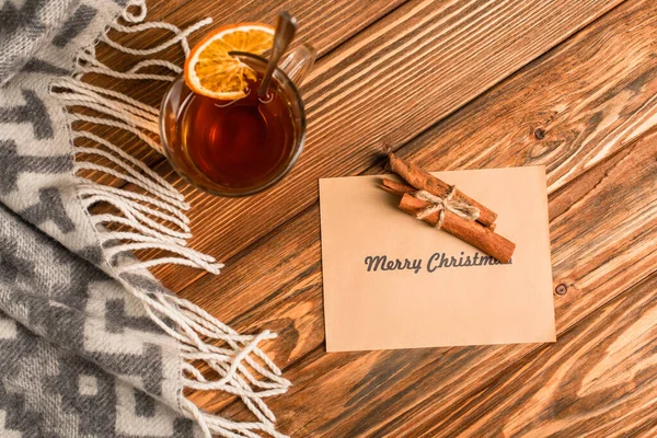 Top view of cup of tea with sliced orange near cinnamon sticks on greeting card with merry christmas lettering on wooden surface — Stock Photo