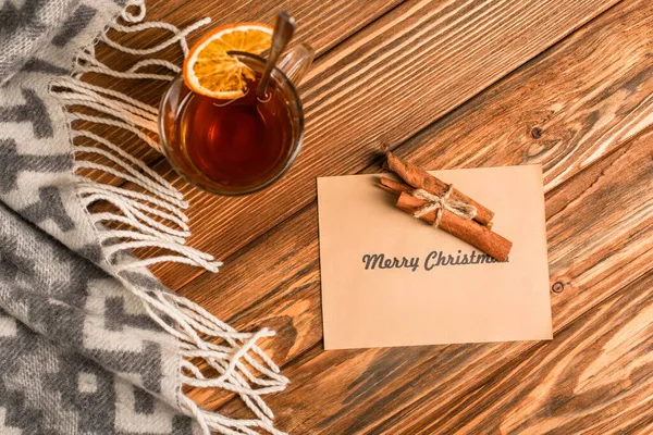 Draufsicht auf Tasse Tee mit geschnittener Orange in der Nähe von Zimtstangen auf Grußkarte mit frohem Weihnachten Schriftzug auf Holzoberfläche — Stockfoto