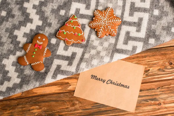 Top view of gingerbread cookies on grey blanket with ornament near greeting card with merry christmas lettering on wooden surface — Stock Photo