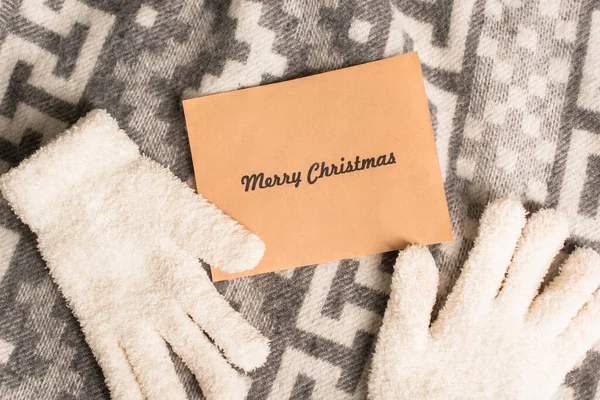 Top view of warm gloves and greeting card with merry christmas lettering on grey blanket with ornament — Stock Photo