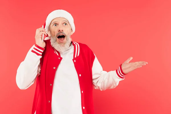 Surprised middle aged man in santa hat pointing with hand while having phone call isolated on red — Stock Photo