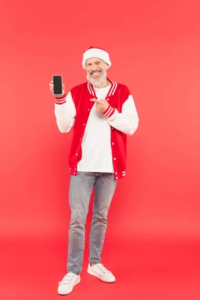 Full length of cheerful middle aged man in santa hat pointing at smartphone with blank screen on red — Stock Photo