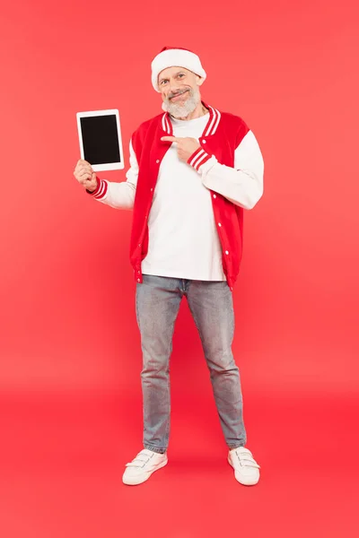 Full length of happy middle aged man in santa hat pointing at digital tablet on red — Stock Photo