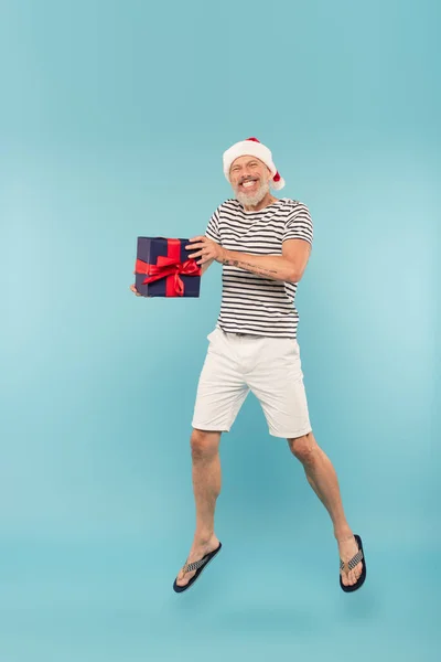 Pleine longueur de heureux homme d'âge moyen en santa chapeau sautant avec boîte cadeau sur bleu — Photo de stock