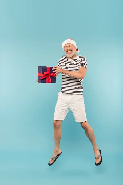 Full length of happy middle aged man in santa hat jumping with gift box on blue — Stock Photo
