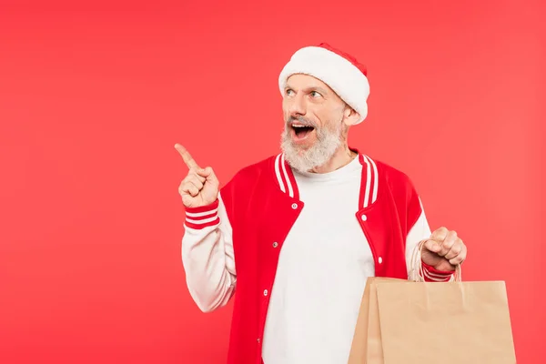 Excité homme d'âge moyen pointant avec le doigt et tenant sac à provisions isolé sur rouge — Photo de stock