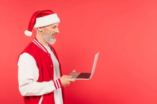 Vue latérale de joyeux pigiste d'âge moyen dans santa chapeau tenant ordinateur portable isolé sur rouge — Photo de stock