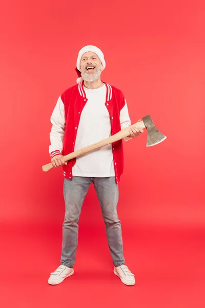 Full length of smiling middle aged man in santa hat holding axe on red — Stock Photo