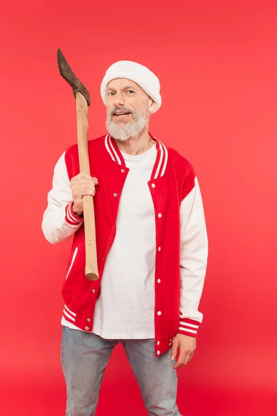 Middle aged man in santa hat holding axe on red — Stock Photo