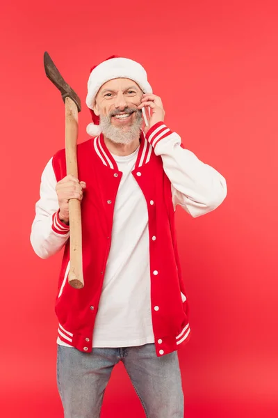 Feliz hombre de mediana edad en sombrero de santa sosteniendo hacha mientras habla en el teléfono inteligente en rojo - foto de stock