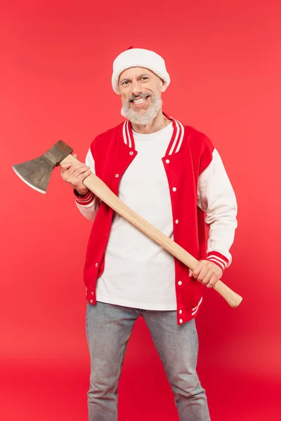 Happy middle aged man in santa hat holding axe on red — Stock Photo