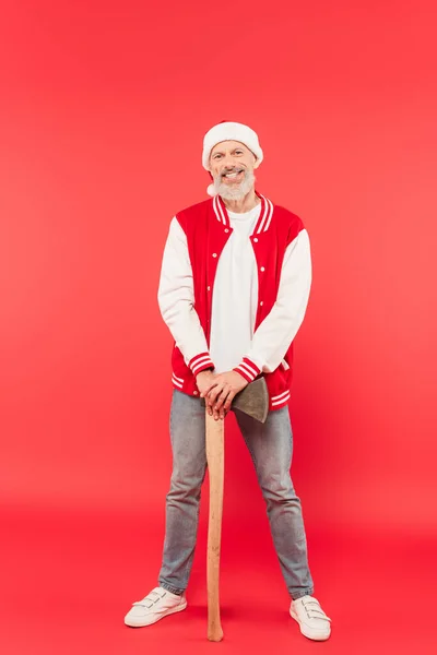 Full length of happy middle aged man in santa hat holding axe while standing on red — Stock Photo