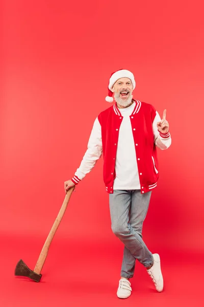 Full length of happy middle aged man in santa hat holding axe and pointing with finger on red — Stock Photo