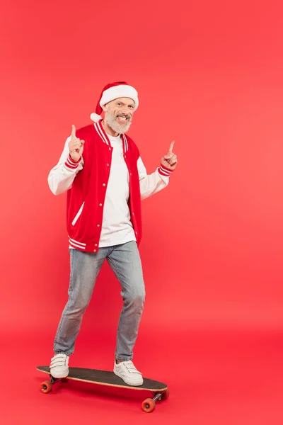 Full length of cheerful middle aged man in santa hat riding skateboard and pointing with fingers on red — Stock Photo