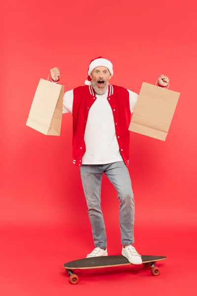 Longitud completa de sorprendido hombre de mediana edad en santa hat celebración de bolsas de compras mientras monta monopatín en rojo - foto de stock
