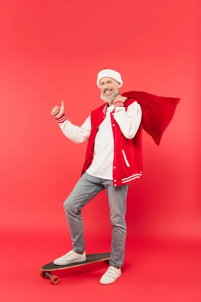 Full length of cheerful middle aged man in hat holding santa sack while riding skateboard and showing thumb up on red — Stock Photo