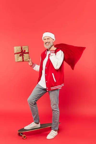 Pleine longueur de gai homme d'âge moyen dans le chapeau tenant santa sac et présent tout en chevauchant planche à roulettes sur rouge — Photo de stock