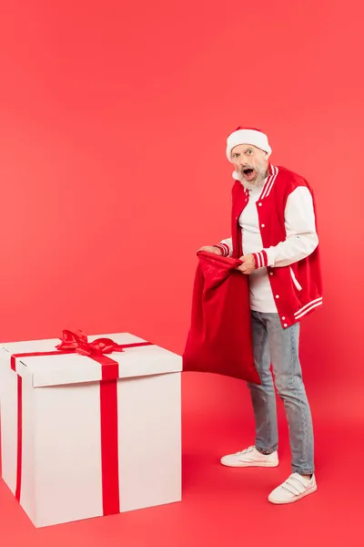 Full length of shocked middle aged man in hat holding santa sack near huge present on red — Stock Photo
