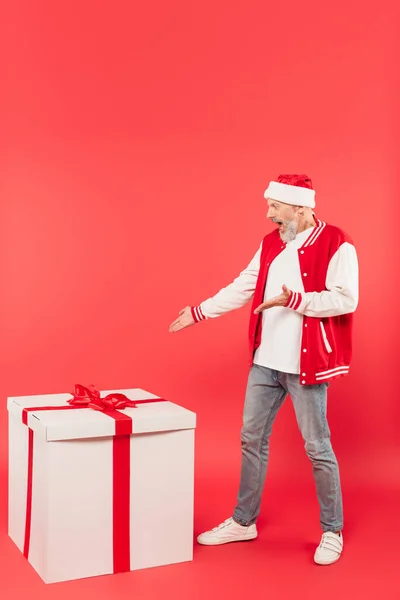 Full length of shocked middle aged man in santa hat pointing at huge present on red — Stock Photo