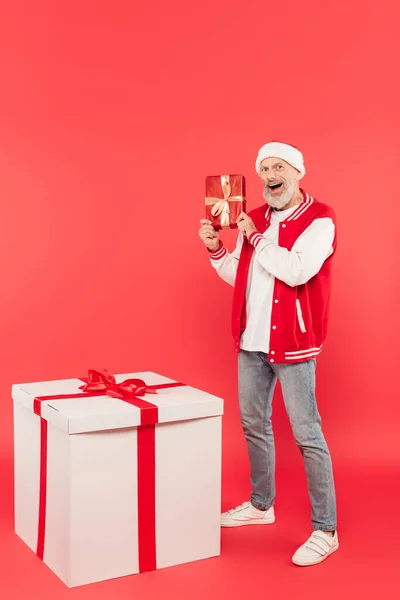Pleine longueur de l'homme d'âge moyen excité dans le chapeau santa tenant boîte cadeau près de cadeau énorme sur rouge — Photo de stock