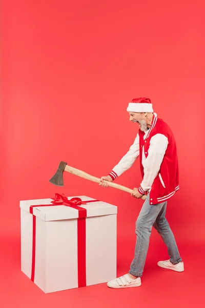 Longitud completa de hombre de mediana edad en santa sombrero sosteniendo hacha mientras grita cerca de regalo enorme en rojo - foto de stock