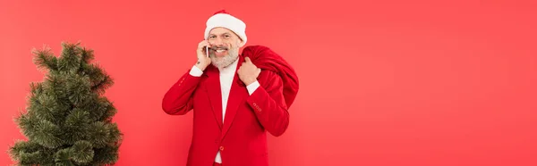 Happy middle aged man in hat and suit holding santa sack and having phone call near pine tree isolated on red, banner — Stock Photo