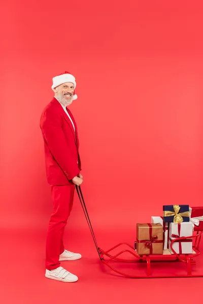 Longitud completa de hombre alegre de mediana edad en sombrero de santa y traje con trineo con regalos en rojo - foto de stock