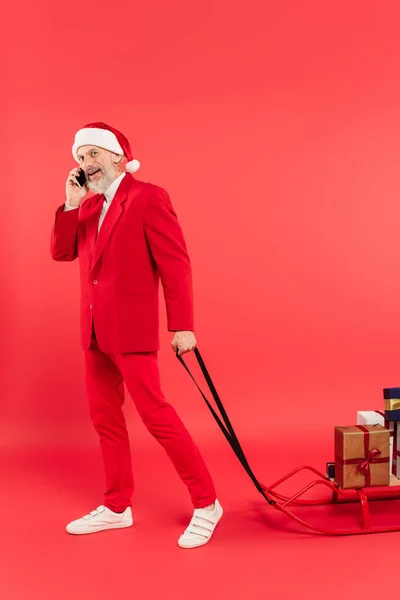 Comprimento total do homem de meia-idade em santa chapéu e terno segurando trenó com presentes enquanto conversa no smartphone em vermelho — Fotografia de Stock