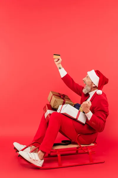 Longitud completa de hombre de mediana edad feliz en sombrero de santa y traje sentado en trineo con regalos mientras sostiene la tarjeta de crédito en rojo - foto de stock