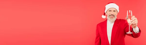 Happy middle aged man in suit and santa hat holding glass of champagne isolated on red, banner — Stock Photo