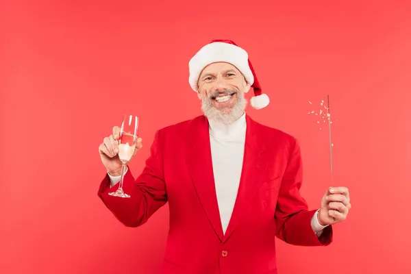 Happy middle aged man in santa hat holding glass of champagne and sparkler isolated on red — Stock Photo