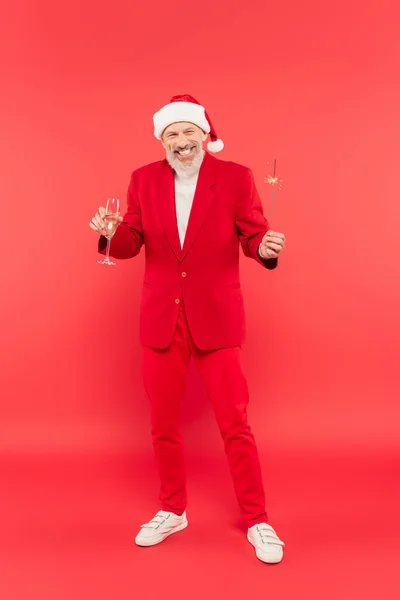 Full length of cheerful middle aged man in suit and santa hat holding glass of champagne and sparkler on red — Stock Photo