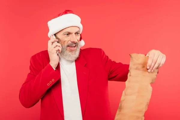 Positivo uomo maturo in Babbo Natale lettera di lettura cappello su carta artigianale mentre si parla su smartphone isolato su rosso — Foto stock