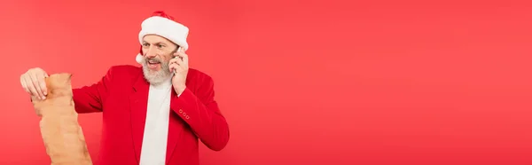 Positive mature man in santa hat reading letter on craft paper while talking on smartphone isolated on red, banner — Stock Photo