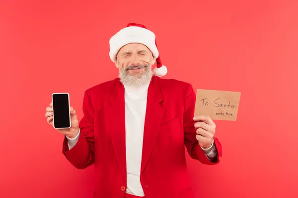 Positive mature man in santa hat holding envelope with lettering and smartphone isolated on red — Stock Photo