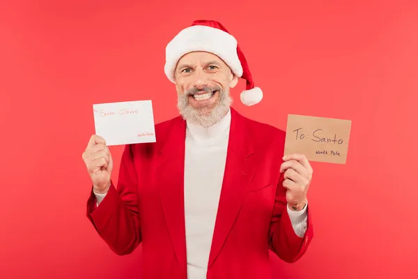 Позитивный зрелый мужчина в шляпе держит конверт и письмо к Санте изолированы на красный — стоковое фото