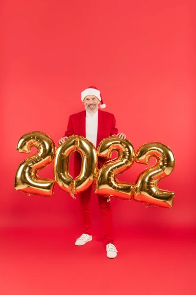 Happy mature man in santa hat standing near 2022 balloons on red — Stock Photo