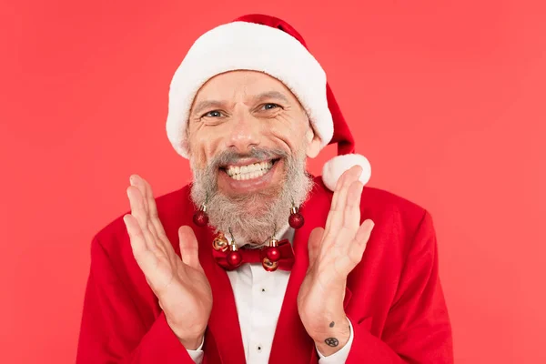 Excité homme mûr dans santa chapeau avec des boules de Noël sur la barbe geste isolé sur rouge — Photo de stock