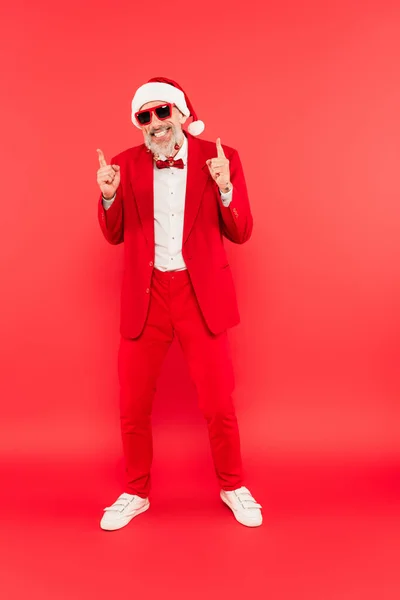 Full length of happy mature man in santa hat and sunglasses with christmas balls on beard pointing up on red — Stock Photo