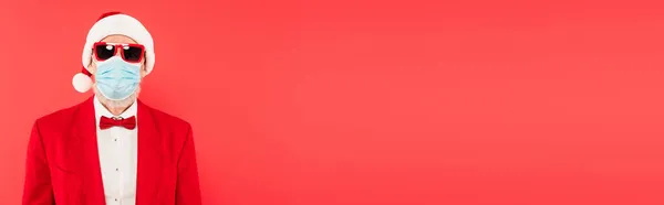 Homem maduro em santa hat, máscara médica e óculos de sol isolados em vermelho, banner — Fotografia de Stock