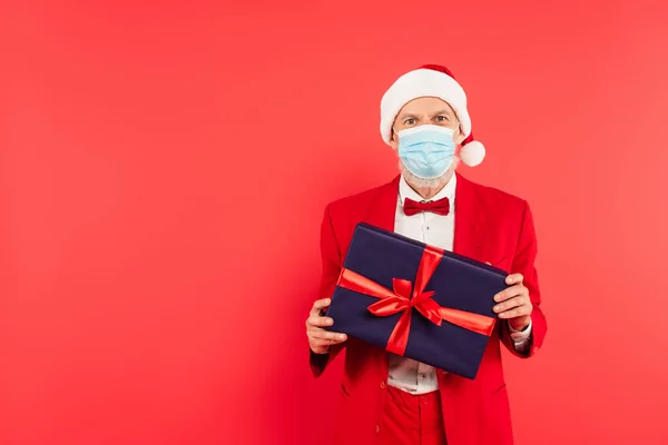 Homem maduro em santa chapéu e máscara médica segurando presente isolado no vermelho — Fotografia de Stock