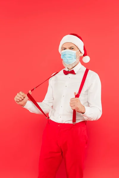 Mature man in santa hat and medical mask adjusting suspenders isolated on red — Stock Photo