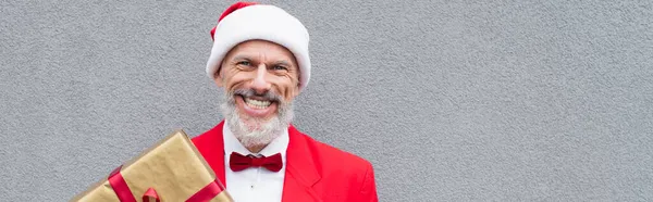 Hombre maduro feliz en sombrero de santa celebración regalo envuelto cerca de la pared gris, bandera - foto de stock