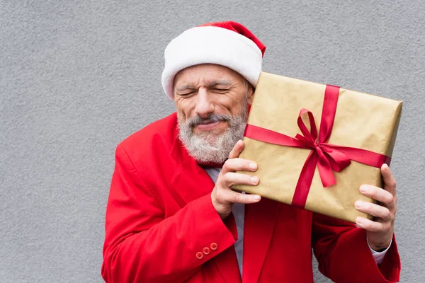 Щасливий зрілий чоловік у капелюсі Санта тримає загорнутий подарунок біля сірої стіни — стокове фото