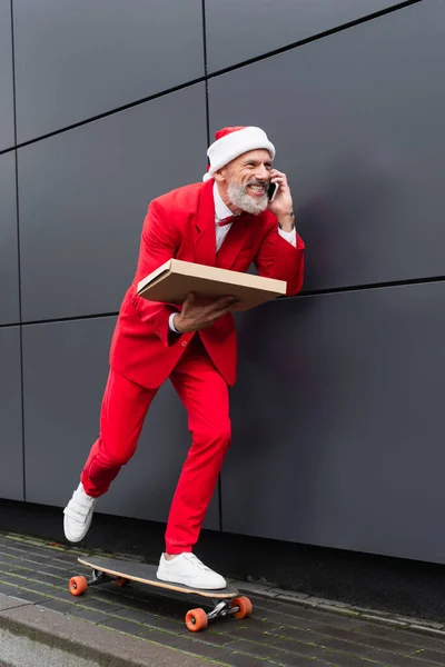 Happy mature man in santa hat holding pizza box and riding on skateboard while talking on cellphone — Stock Photo