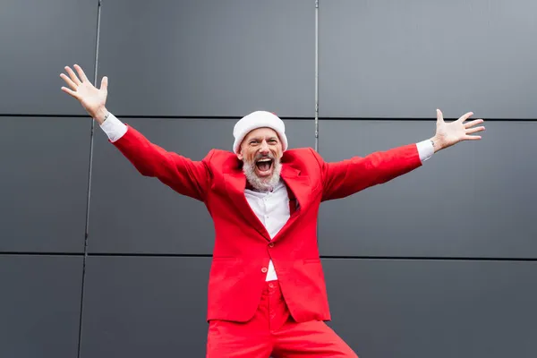 Excité homme mature dans santa chapeau debout avec les mains tendues près du mur gris — Photo de stock